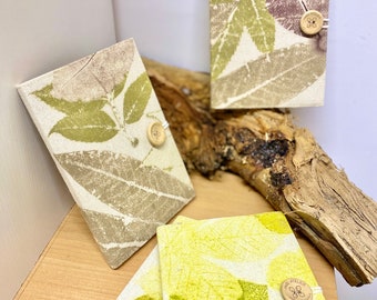 May include: Three handmade notebooks with leaf prints on the covers. The covers are a light beige color with green and brown leaf prints. Each notebook has a wooden button closure with a small wooden tag that says "L'Atelier" on it.