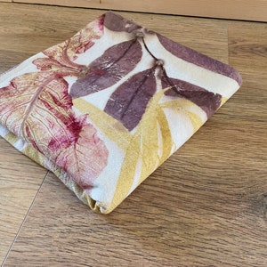May include: A folded fabric with a white background and a leaf pattern in shades of yellow, red, and purple.