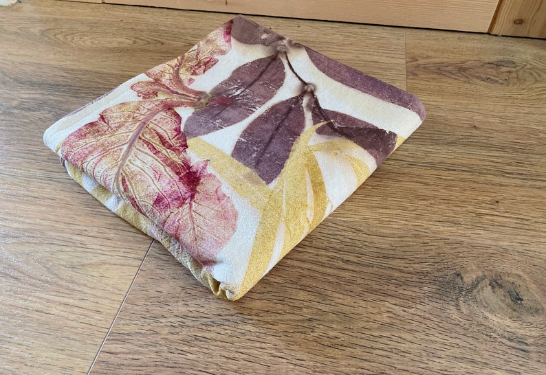 May include: A folded fabric with a white background and a leaf pattern in shades of yellow, red, and purple.