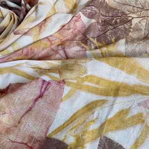 May include: A white fabric with a natural leaf print in shades of brown, yellow, and pink. The fabric is folded and draped, showing the intricate details of the leaf patterns.