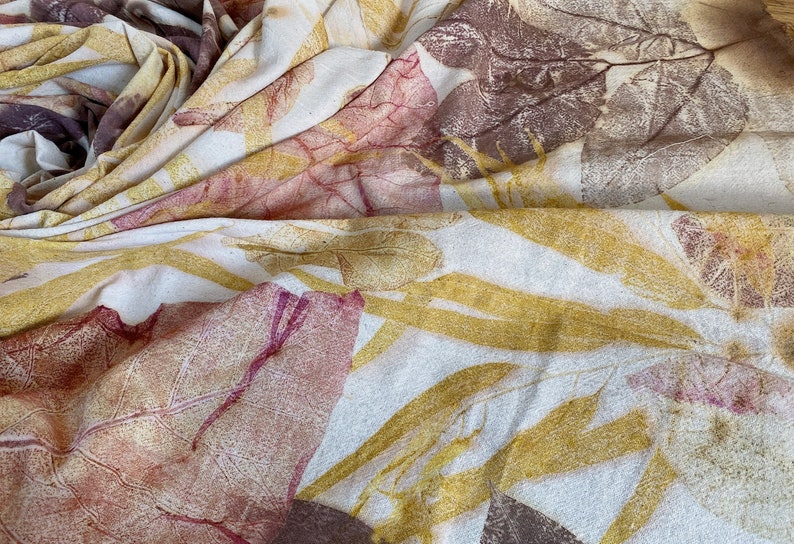 May include: A white fabric with a natural leaf print in shades of brown, yellow, and pink. The fabric is folded and draped, showing the intricate details of the leaf patterns.