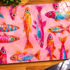 May include: A rectangular glass chopping board featuring a pink backdrop and vibrant fish illustrations. The fish are depicted in pink, orange, and blue hues. A knife and fresh produce are visible in the background.