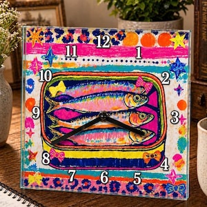 Puede incluir: Reloj de pared cuadrado con un diseño vibrante y colorido. La esfera del reloj presenta una ilustración de una lata de sardinas con tres peces, sobre un fondo rosa, azul y amarillo. Las manecillas del reloj son negras.