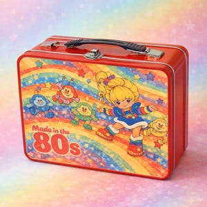 Może przedstawiać: Czerwony metalowy lunchbox z kolorową ilustracją blond dziewczynki i postaci z kreskówek na tle tęczy. Widoczny jest napis "Made in the 80s". Posiada czarny uchwyt i srebrne zapięcie.