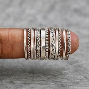 May include: A set of eleven silver rings arranged in a row, featuring various textures and patterns including twisted rope, smooth bands, floral engravings, and geometric cross-hatch designs, all crafted from polished silver metal with oxidised details.