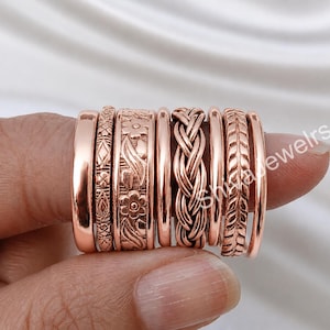 May include: A set of eight rose gold-toned rings featuring various textures and patterns, including a smooth band, a geometric etched band, a floral embossed band, a braided design, and a leaf-patterned band, all displayed together on a finger.