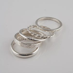 May include: A collection of five silver rings. The rings have various designs, including a smooth band, a textured band, and a braided band. The rings are displayed on a white surface.