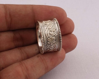 Könnte beinhalten: Ein breiter, silberfarbener Ring mit einem aufwendigen Blumen- und Blättermuster. Der Ring hat eine strukturierte Oberfläche mit einem Perlenrand. Das Design umfasst eine zentrale Blume und wirbelndes Laub.