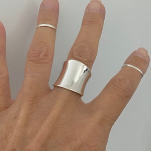 May include: A large, polished silver ring with a curved design is worn on a finger. Two other thin silver rings are also visible on adjacent fingers. The jewellery is displayed against a neutral background.