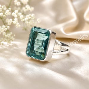 Pode incluir: Um anel de prata com uma grande pedra preciosa retangular facetada de cor azul-clara, engastada num aro de metal liso. A banda do anel divide-se em duas secções distintas que se ligam ao engaste principal, criando um design aberto.