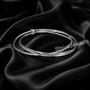 May include: Three silver bangles with a textured design and a knot closure, resting on a black satin surface. The bangles are intertwined, creating a layered effect. The text "Shivajewelrs" is visible.