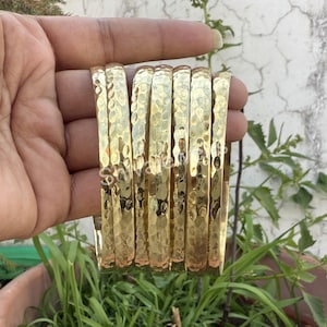 May include: A set of seven gold-toned hammered metal bangles. The bangles are stacked on top of each other and are being held in a hand.