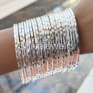 Pode incluir: Um conjunto de pulseiras de prata martelada. As pulseiras estão empilhadas no pulso de uma pessoa.