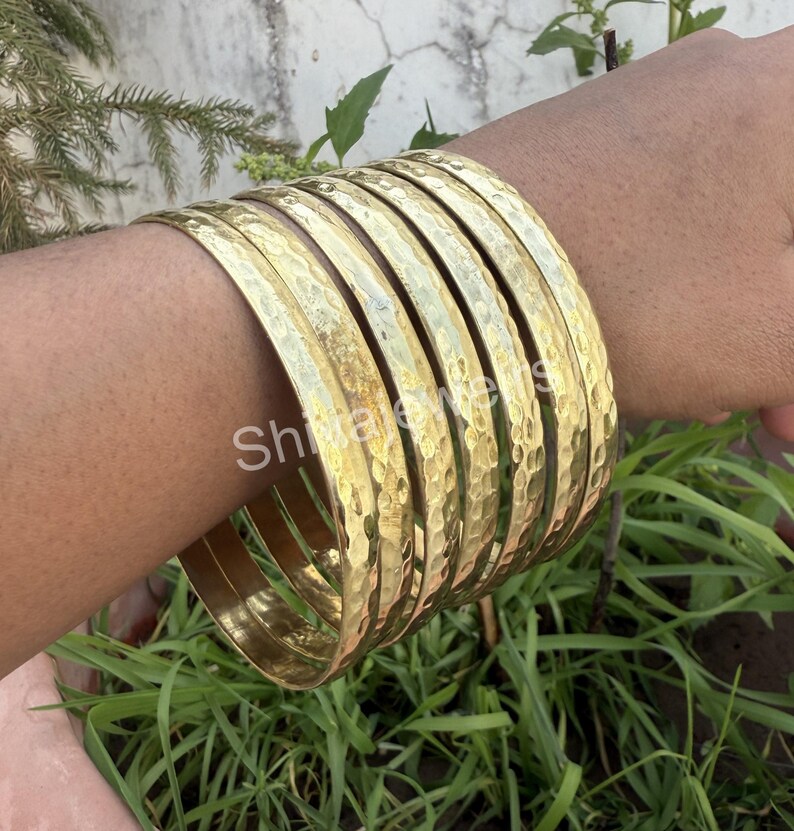 May include: A set of eight gold-toned hammered metal bangles. The bangles are stacked on a person's arm.