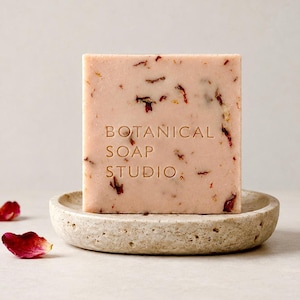 May include: A square, light pink bar of soap embedded with dried flower petals, featuring the embossed text "BOTANICAL SOAP STUDIO" on the front, resting on a round, textured, beige stone dish surrounded by scattered red rose petals.