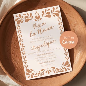 Op de afbeelding: Een uitnodiging voor een bruidsdouche met de tekst "Viva la Novia" en "Angelique" in elegant schrift. De uitnodiging heeft een bloemenrand en ligt op een terracotta bord. De tekst "Edit in Canva" is ook zichtbaar.