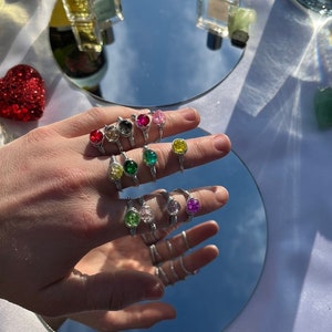 May include: A hand wearing a variety of silver rings with colorful glass stones. The rings are stacked on each finger. The stones are green, pink, yellow, clear, and purple.