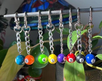 May include: Eight silver keychains with different coloured billiard balls attached. The balls are numbered 1 through 8 and are green, yellow, purple, red, black, and blue.