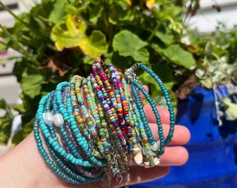 May include: A stack of colorful beaded bracelets in shades of turquoise, green, blue, red, yellow, and gold. The bracelets are layered on top of each other and feature small white beads.