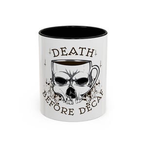 May include: White ceramic coffee mug with a black interior. The mug features a skull design with the words "DEATH BEFORE DECAF" printed around the skull. The skull is designed to look like a coffee cup.