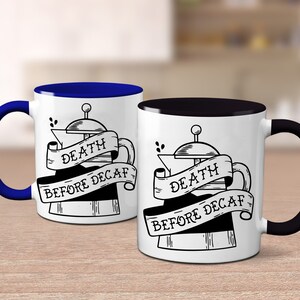 May include: Two white coffee mugs with black and blue handles. Each mug features a black and white illustration of a coffee maker with a banner that reads "DEATH BEFORE DECAF".