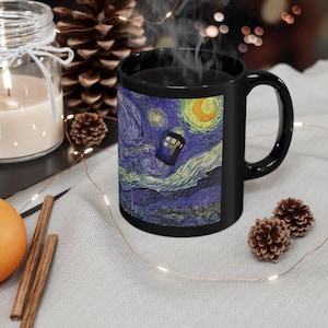 May include: Black ceramic mug with a print of Van Gogh's "Starry Night" and a TARDIS. The mug has a curved handle and is filled with a steaming dark liquid. The mug is surrounded by decorative elements.