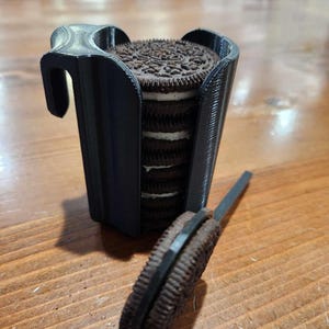 May include: A black 3D printed cookie holder with a stack of Oreo cookies inside. A single Oreo cookie is lying on the table next to the holder.