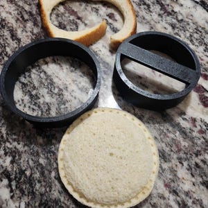 May include: Three round bread molds, two are black plastic and one is white bread. The bread molds are on a speckled countertop.