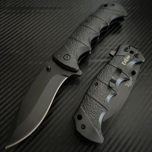 May include: Two black folding knives with textured handles. The larger knife has a curved blade and the smaller knife has a straight blade. Both knives have the text "Edge-Up" printed on them.