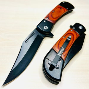 May include: Two black folding pocket knives with wooden handles. The knives have a black blade and a silver clip. The knives are on a light brown wooden surface. The text "Elite Edge" is printed on the handle of one of the knives.