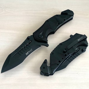 May include: Two black folding knives with a serrated blade and a safety feature. The knives have a black handle with a textured grip and the words "MTech USA" printed on the side.