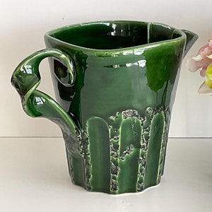 May include: A green ceramic jug with a unique, sculpted handle. The jug has a textured, slightly rough surface.