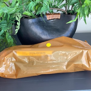 K&ouml;nnte beinhalten: Eine orange-braune Verpackung mit einem gelben Aufkleber befindet sich vor einer schwarzen dekorativen Schale mit gr&uuml;nen Pflanzen. Auf der Verpackung steht der Text "312A/CF382A". Der Hintergrund ist eine dunkelgraue Oberfl&auml;che.