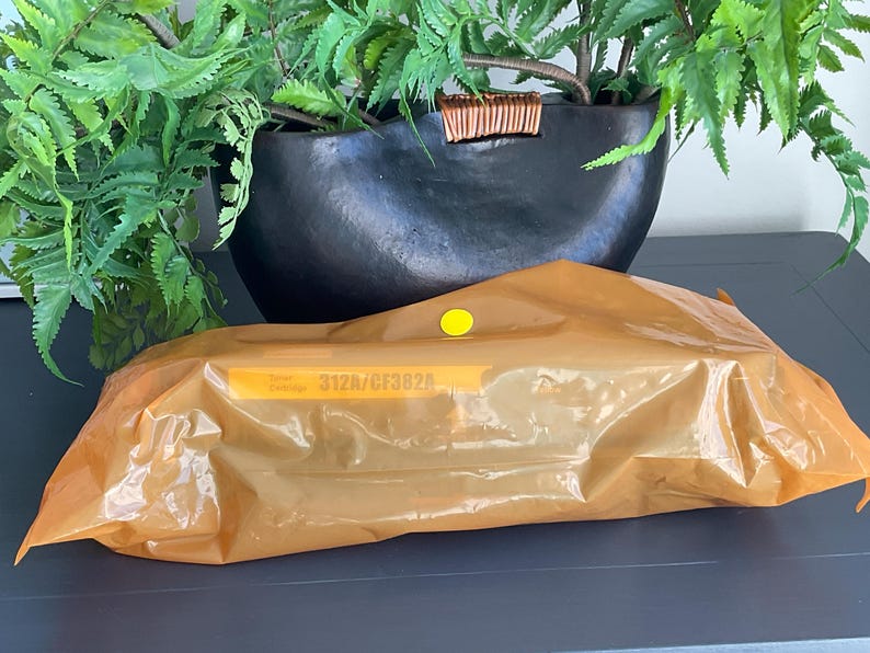 K&ouml;nnte beinhalten: Eine orange-braune Verpackung mit einem gelben Aufkleber befindet sich vor einer schwarzen dekorativen Schale mit gr&uuml;nen Pflanzen. Auf der Verpackung steht der Text "312A/CF382A". Der Hintergrund ist eine dunkelgraue Oberfl&auml;che.