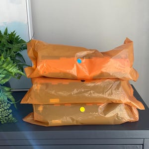 K&ouml;nnte beinhalten: Drei orangefarbene Pakete sind auf einer dunkelgrauen Oberfl&auml;che gestapelt. Die Pakete sind in durchscheinendes orangefarbenes Plastik verpackt. Der Hintergrund umfasst einen Farn und ein gerahmtes Kunstwerk.