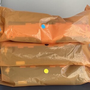 K&ouml;nnte beinhalten: Drei Pakete, verpackt in orangefarbenem, durchscheinendem Kunststoff, sind &uuml;bereinander gestapelt. Die Pakete haben orangefarbene und braune Elemente. Jedes Paket hat einen farbigen Aufkleber: blau, schwarz und gelb. Die Pakete liegen auf einer dunklen Oberfl&auml;che.