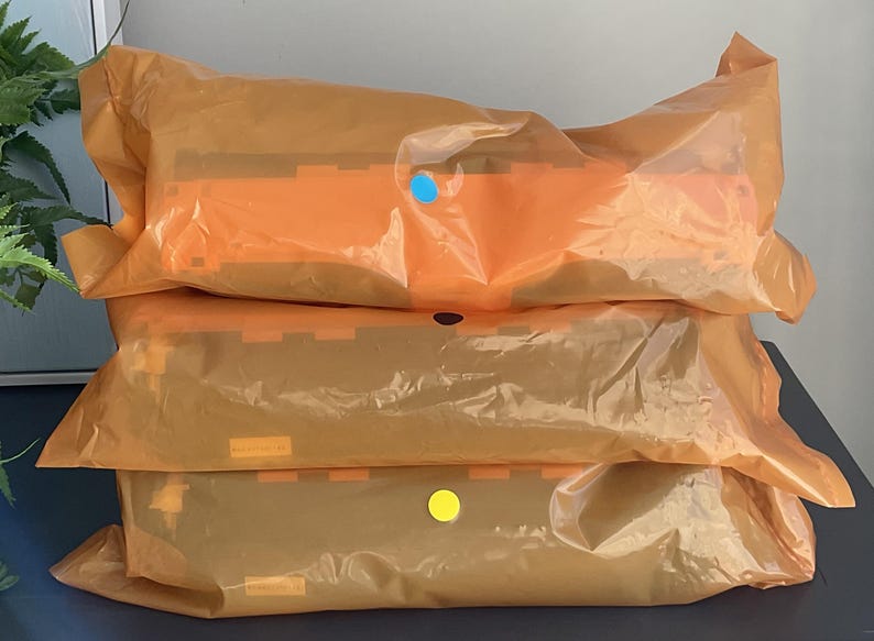 K&ouml;nnte beinhalten: Drei Pakete, verpackt in orangefarbenem, durchscheinendem Kunststoff, sind &uuml;bereinander gestapelt. Die Pakete haben orangefarbene und braune Elemente. Jedes Paket hat einen farbigen Aufkleber: blau, schwarz und gelb. Die Pakete liegen auf einer dunklen Oberfl&auml;che.