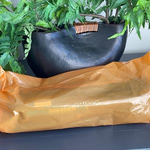 K&ouml;nnte beinhalten: Eine orangefarbene Plastikverpackung liegt auf einer dunklen Oberfl&auml;che, mit einem schwarzen, oval geformten Pflanzgef&auml;&szlig; und gr&uuml;nen Bl&auml;ttern im Hintergrund. Die Verpackung hat Text aufgedruckt. Der Pflanztopf hat einen dekorativen Holzakzent.