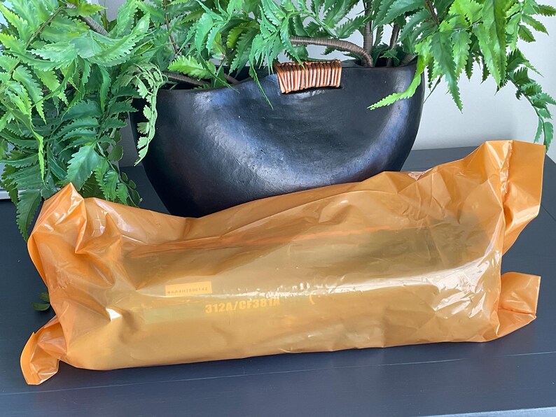 K&ouml;nnte beinhalten: Eine orangefarbene Plastikverpackung liegt auf einer dunklen Oberfl&auml;che, mit einem schwarzen, oval geformten Pflanzgef&auml;&szlig; und gr&uuml;nen Bl&auml;ttern im Hintergrund. Die Verpackung hat Text aufgedruckt. Der Pflanztopf hat einen dekorativen Holzakzent.