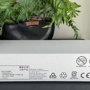 May include: A white HP printer box with text in multiple languages, including Japanese and Chinese. The box has various symbols and recycling information. A black planter with green plants is in the background.