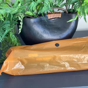 K&ouml;nnte beinhalten: Ein schwarzer, oval geformter Pflanztopf mit strukturierter Oberfl&auml;che, gef&uuml;llt mit gr&uuml;nen Bl&auml;ttern. Eine orangefarbene Plastikverpackung mit einem kleinen Loch befindet sich im Vordergrund. Die Verpackung hat Text aufgedruckt.