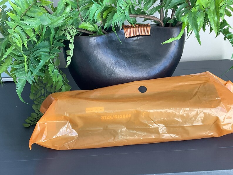 K&ouml;nnte beinhalten: Ein schwarzer, oval geformter Pflanztopf mit strukturierter Oberfl&auml;che, gef&uuml;llt mit gr&uuml;nen Bl&auml;ttern. Eine orangefarbene Plastikverpackung mit einem kleinen Loch befindet sich im Vordergrund. Die Verpackung hat Text aufgedruckt.