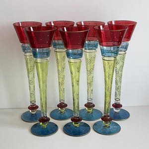 May include: A set of eight vintage champagne flutes. Each glass features a red bowl with white dots, a blue band, a yellow stem with gold swirls, and a blue base. The glasses have a gold rim.