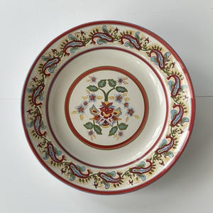 May include: A white ceramic dinner plate with a red, yellow and blue floral design. The plate has a paisley border.
