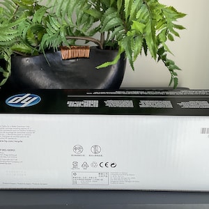 May include: A white HP printer cartridge box with black accents, featuring the HP logo and product information. The box is rectangular and sits on a dark surface, with green foliage in the background.