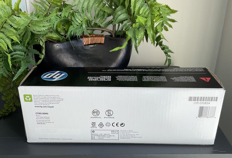 May include: A white HP printer cartridge box with black accents, featuring the HP logo and product information. The box is rectangular and sits on a dark surface, with green foliage in the background.