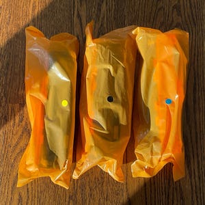 K&ouml;nnte beinhalten: Drei Artikel, verpackt in durchscheinendem orangefarbenem Kunststoff, jeweils mit einem unterschiedlich farbigen Punkt: gelb, schwarz und blau. Die Artikel liegen auf einer Holzoberfl&auml;che mit Schatten.