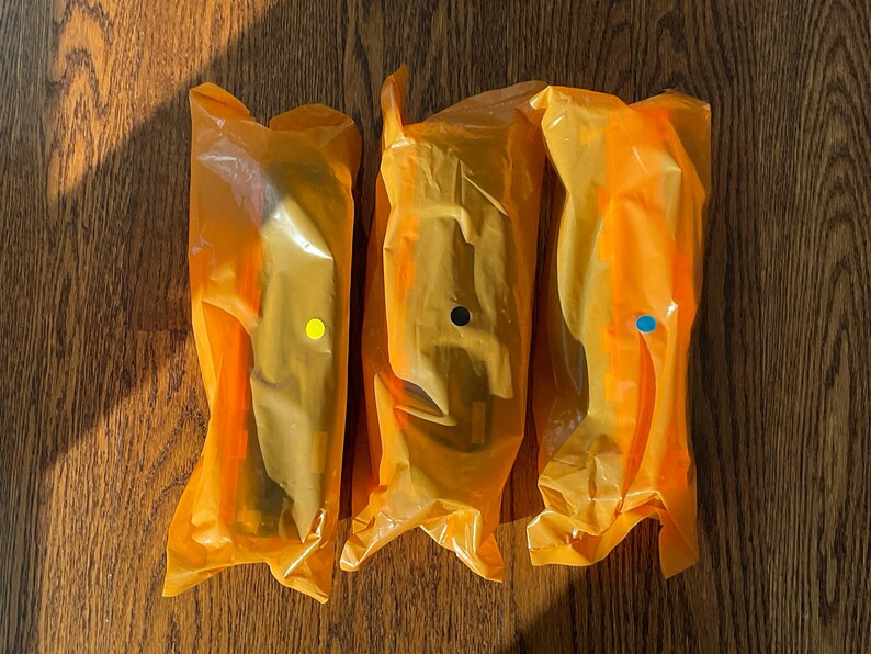 K&ouml;nnte beinhalten: Drei Artikel, verpackt in durchscheinendem orangefarbenem Kunststoff, jeweils mit einem unterschiedlich farbigen Punkt: gelb, schwarz und blau. Die Artikel liegen auf einer Holzoberfl&auml;che mit Schatten.