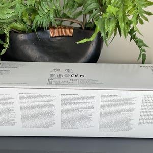 May include: A white rectangular box with printed text and a barcode. The box is sitting on a dark surface, with a green plant and a black pot in the background.