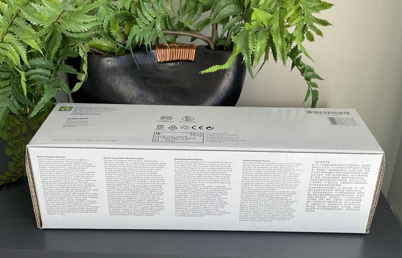 May include: A white rectangular box with printed text and a barcode. The box is sitting on a dark surface, with a green plant and a black pot in the background.